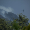 Helicóptero con dispositivo para descarga de agua sobre una zona montañosa con presencia de humo, mientras se realizan labores de atención a un incendio forestal entre áreas boscosas.