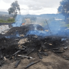Carbón vegetal, cenizas y residuos de madera humeantes en un área intervenida al aire libre, con sacos apilados y vegetación alrededor en una zona rural.