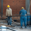 Obreros con cascos de seguridad trabajan en una obra de construcción, manipulando tuberías y equipos frente a una estructura de ladrillo.