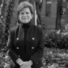 Retrato en blanco y negro de Liliana Caballero Durán de pie en un jardín con árboles y vegetación al fondo, vistiendo chaqueta oscura de doble botonadura y cuello alto.