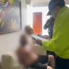 Persona con el rostro difuminado sentada en un sofá mientras otra persona con chaqueta reflectiva amarilla permanece de pie a su lado en una sala con cuadros en la pared