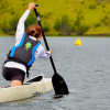 Persona practicando canotaje en un lago rodeado de vegetación, mientras impulsa una embarcación con un remo sobre el agua.