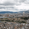 Vista panorámica de Bogotá con áreas residenciales y comerciales, edificios de diferentes alturas, vías urbanas y montañas al fondo bajo un cielo cubierto de nubes grises.