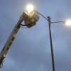 Poste de alumbrado público encendido mientras una plataforma elevadora realiza labores de mantenimiento en la parte superior, bajo un cielo nublado al atardecer.