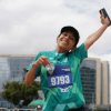 Participante de la Bimbo Global Race con camiseta verde y dorsal 9193 levanta un brazo mientras corre. Al fondo se observan edificaciones y el cielo despejado.
