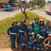 Cuadrilla de jardinería posa en una jardinera urbana junto a plantas ornamentales y árboles jóvenes; al fondo se observan una vía, vehículos y edificaciones.