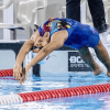 Nadador realiza la partida de espalda desde el borde de una piscina de competencia. La deportista viste traje de baño azul, gafas y gorro de natación, mientras se arquea hacia atrás para ingresar al agua.