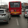 Bus articulado rojo de TransMilenio circula por una vía urbana junto a un automóvil gris y una motocicleta con carga en la parte trasera.