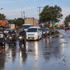 Motocicletas, vehículos particulares y camiones circulan por una vía mojada tras una lluvia, con reflejos sobre el pavimento y tránsito lento en ambos sentidos. Al fondo se observan árboles, postes de energía y señalización vial.