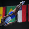 Deportista del Equipo Bogotá ejecuta un salto en competencia de atletismo, captado en el aire durante la prueba. Al fondo se observan banderas de distintos países participantes.