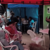 Personas sentadas en sillas plásticas asisten a una charla en un espacio cubierto, mientras una persona expone junto a una mesa.