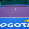 Cancha de tenis con superficie morada y zonas laterales azules, delimitada por líneas blancas y una red en el centro. En el primer plano se observa el logo de Bogotá en color blanco y amarillo sobre la superficie de la cancha.