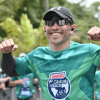 Participante de una carrera atlética viste camiseta verde de la Bimbo Global Race, gorra negra, gafas oscuras y audífonos inalámbricos, mientras avanza por el recorrido junto a otros corredores en un entorno al aire libre rodeado de árboles.