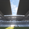 Vista interior del nuevo estadio El Campín de Bogotá con graderías, cancha de césped en el centro y una gran cubierta metálica parcialmente abierta que deja ver el cielo. La luz del sol ingresa por la apertura del techo e ilumina una franja del campo de juego.