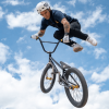 Luis Oswaldo Rincón ejecuta una maniobra aérea en una bicicleta BMX durante una competencia de ciclismo extremo. La bicicleta se encuentra inclinada en el aire y el deportista viste casco, camiseta oscura y pantalón claro.