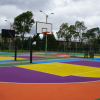 Cancha deportiva multicolor con tablero y aro de baloncesto, rodeada por cerramiento metálico, árboles y cielo nublado al fondo.