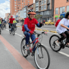 Ciclistas y peatones transitan por una vía urbana durante una jornada recreativa. En primer plano, dos personas montan bicicleta con casco mientras otras personas avanzan en bicicleta y caminando por la calzada.