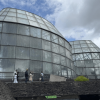 Vista exterior de los domos de vidrio del Tropicario del Jardín Botánico de Bogotá. En primer plano se observa un sendero elevado por el que transitan varias personas, mientras al fondo se aprecia un cielo parcialmente nublado y vegetación alrededor de la estructura.
