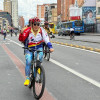 Persona con camiseta blanca, roja y azul monta una bicicleta sobre una vía de la Ciclovía de Bogotá; al fondo se observan edificaciones, vehículos y otros ciclistas.