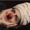 Imagen de la cara de una mujer caracterisada como payasa, con una nariz redonda roja y un sombrero blanco.
