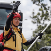 Persona con casco rojo, guantes y uniforme amarillo sostiene una paleta de señalización con la palabra “SIGA” mientras levanta una mano.