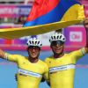 Dos deportistas de patinaje de velocidad con uniformes amarillos de Colombia y cascos blancos sostienen una bandera de Colombia sobre sus cabezas en una pista de competencia.
