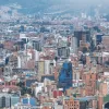 Vista panorámica del centro de Bogotá con numerosos edificios de diferentes alturas y colores, vías urbanas y montañas al fondo parcialmente cubiertas por neblina.