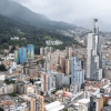 Vista panorámica de Bogotá con edificios de gran altura y zonas residenciales, junto a los cerros cubiertos de vegetación y neblina al fondo bajo un cielo nublado.