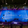 Vista aérea de varias canchas de pádel iluminadas en color azul dentro de un complejo deportivo cubierto, con jugadores en acción y áreas de circulación alrededor de los escenarios deportivos.