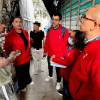 Personal con chaquetas rojas realiza una visita de inspección en una bodega de alimentos. Varias personas conversan entre pilas de sacos almacenados y un camión de carga estacionado junto al área de trabajo.