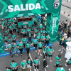 Cientos de participantes cruzan el arco de salida de la Bimbo Global Race 2026 sobre una vía urbana. El pórtico verde muestra el texto “SALIDA”, los logotipos de Bimbo Global Race 2025 y la frase “COMPARTIMOS LO BUENO”. A los lados hay espectadores detrás de vallas, mientras corredores con camisetas verdes avanzan por la línea de partida.