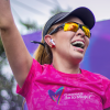 Participante de la Carrera de la Mujer con camiseta rosada, gorra negra, gafas deportivas y audífonos inalámbricos, levantando un brazo durante una actividad del evento al aire libre.