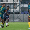 Jugador controla un balón durante un partido de fútbol en una cancha de césped, mientras otros participantes se desplazan a su alrededor. Al fondo se observa una cerca metálica y estructuras deportivas.