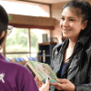 Imagen de una mujer sonriendo mientras entrega un libro a funcionaria de BibloRed de espaldas.