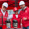 Alcalde Mayor de Bogotá, Carlos Fernando Galán, junto a Leonidas Narváez, gerente de la Línea 1 del Metro de Bogotá y Neida Ospina, vendedora de café en el carro ambulante 'Metro Tinto' visten con chalecos y cascos de seguridad rojos y blancos permanecen bajo una carpa roja junto a una mesa con termos de café. En las prendas se observan los logos de Bogotá y Metro de Bogotá.