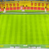 Vista general de la cancha del estadio El Campín con césped verde cortado en franjas longitudinales. En la parte superior se observan graderías con sillas amarillas y rojas, una franja publicitaria del IDRD y parte de la cubierta del escenario deportivo.