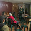 Charla informativa sobre el cuidado de animales de compañía en un salón comunitario. Varias personas sentadas en sillas de plástico observan una presentación proyectada en pantalla, mientras una persona expone al frente y un agente de policía permanece de pie al lateral con proyección en pared de un perro en zona verde.