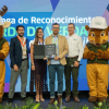 Entrega de reconocimiento ambiental a una entidad distrital. En una tarima, varias personas sostienen un galardón mientras posan para una fotografía, acompañadas por una mascota institucional. Al fondo se observa una pantalla azul con texto relacionado con el programa "Sello Verde de Verdad".