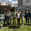 Grupo de personas reunidas alrededor de un árbol recién sembrado en una zona verde de un parque. Varias personas sostienen herramientas de jardinería mientras, al fondo, se observan edificaciones residenciales y montañas bajo un cielo parcialmente nublado.