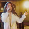 Imagen de una mujer joven de cabello rubio largo cantando con un micrófono.
