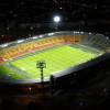 Vista nocturna del estadio El Campín iluminado durante un partido de fútbol, con las tribunas visibles alrededor del terreno de juego y las luminarias encendidas sobre la cancha.