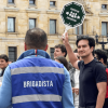 Grupo de personas reunidas en una plaza frente a un edificio institucional. En primer plano, una persona con chaleco azul que dice “BRIGADISTA” observa a otra que sostiene una señal octagonal con el texto “PUNTO DE ENCUENTRO”.