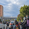 Motociclistas avanzan entre vehículos en una vía de Bogotá durante el día. A un costado de la carretera se observan árboles y, al fondo, edificios residenciales bajo un cielo parcialmente nublado.