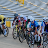 Grupo de ciclistas compite en una pista inclinada de velódromo durante una prueba de ciclismo. Las y los deportistas avanzan en fila sobre bicicletas de ruta, con uniformes de distintos colores y cascos de competición, mientras recorren la curva de la pista de madera.