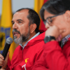 Primer plano de una mesa de reunión institucional. El director del IDRD, Daniel García Cañón, con chaqueta roja sostiene un micrófono y habla, mientras otra persona, también con chaqueta roja, aparece desenfocada en primer plano durante rueda de prensa para explicar situación de la gramilla del estadio El Campín de Bogotá.