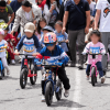 Niñas y niños participan en una carrera de bicicletas sin pedales en una vía urbana, acompañados por adultos y usando cascos de protección.