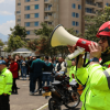 Persona con casco rojo y chaqueta reflectiva usa un megáfono para orientar a un grupo de ciudadanos durante un ejercicio de simulación de emergencia por sismo y otras emergencias en un espacio urbano de Bogotá.