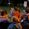 Grupo de personas reunidas en un parque durante una jornada nocturna de preparación para simulacro. Un integrante del cuerpo de bomberos brinda indicaciones mientras una persona sostiene un letrero con la identificación "Edificio B5".