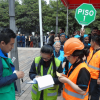 Personas participan en un ejercicio de evacuación en un espacio abierto. En primer plano, varios integrantes de brigadas de emergencia con chalecos reflectivos verdes y naranjas registran información en formularios. Detrás, un grupo organizado de participantes se reúne junto a una señal octagonal verde con el texto “PISO 1”.