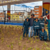 Personal del Jardín Botánico de Bogotá posa junto a jardineras con plantas ornamentales en un espacio público, con una cancha de pádel y edificaciones al fondo.